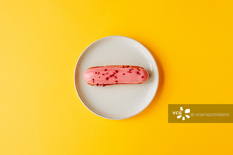 简约的餐桌布置，白色盘子中央放着一块粉色糖霜的泡芙，背景为明亮的黄色。图片素材