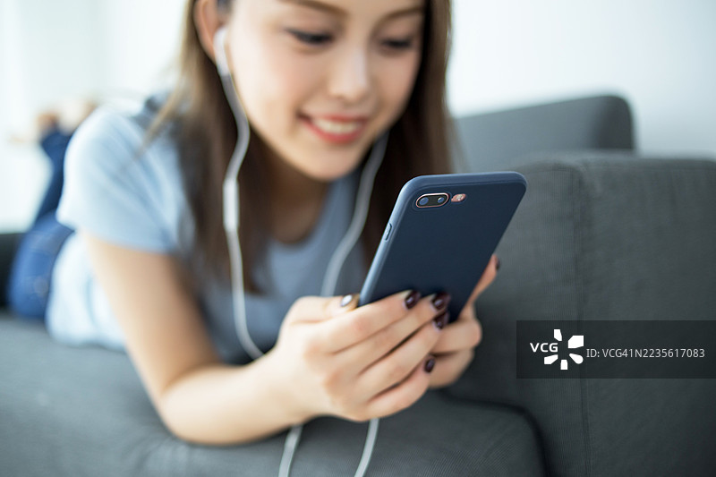 年轻女性躺在沙发上，戴着耳机使用智能手机图片素材