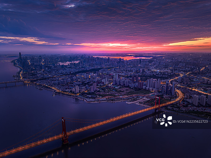黄昏时分，河上横跨一座灯光璀璨的桥，背景是生机勃勃的城市天际线和戏剧性的落日天空。图片素材
