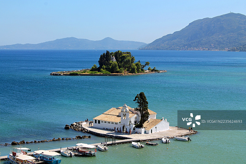 高角度俯瞰一座小岛，上面有一座修道院，远处是一座更大的岛屿，四周被蔚蓝的大海环绕，天空晴朗，希腊，科孚岛图片素材
