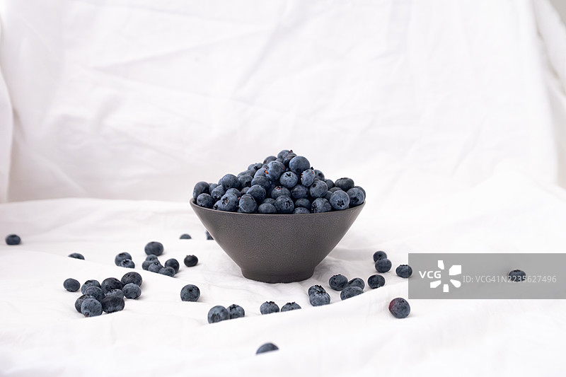 新鲜蓝莓放在深色碗中，周围散落着一些，背景为白色。图片素材