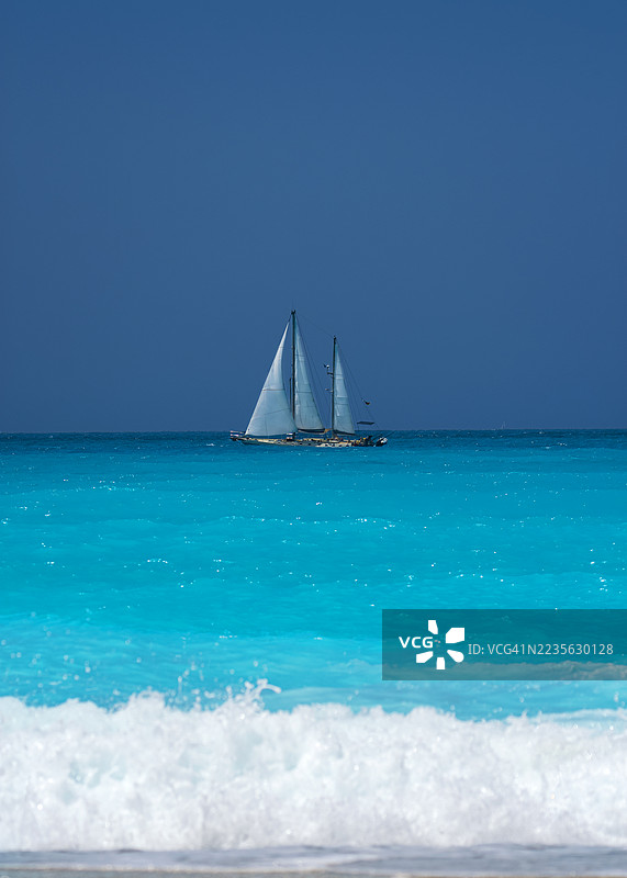 一艘帆船在清澈的蓝天下，滑过生机勃勃的碧绿色海洋，前景中有泡沫翻滚的海浪，希腊，莱夫卡达。图片素材
