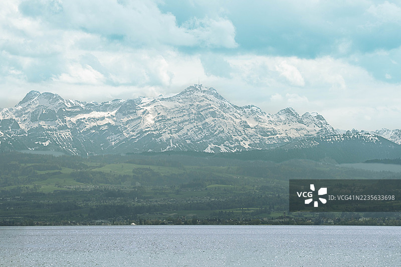 白雪皑皑的雄伟山脉在阴云密布的天空下，高耸于宁静的湖泊之上，展示出一派令人惊叹的自然风光，德国，腓特烈港图片素材