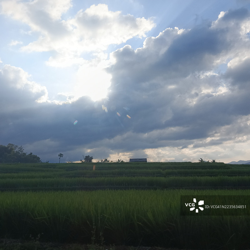 郁郁葱葱的农业田野在戏剧性的天空下，阳光透过云层洒下。图片素材