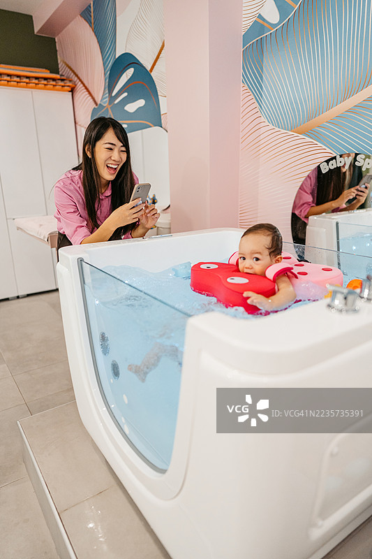 中年母亲用手机拍摄她的女婴在婴儿水疗中心的浴缸里游泳，旁边有浮筒。图片素材