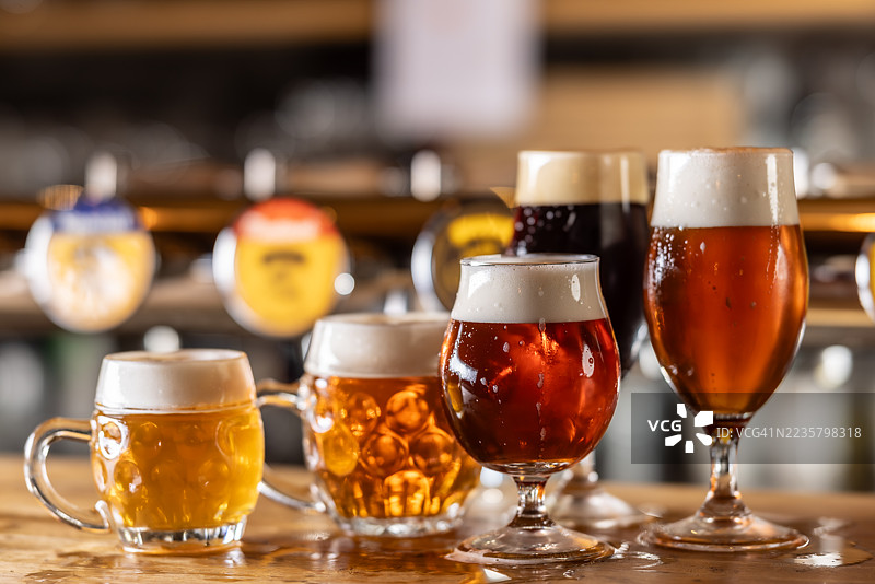 几种不同类型和颜色的生啤酒，盛在不同的酒杯中，摆放在酒吧的木质吧台上。图片素材