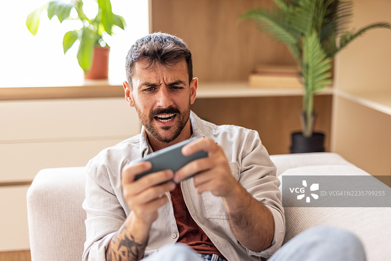 沮丧的男人在家沙发上玩手机游戏失败图片素材