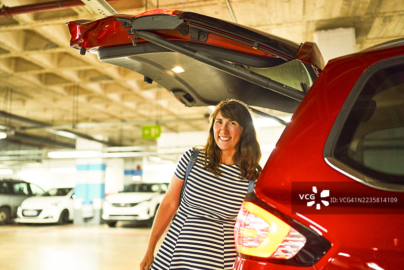 中年女性在停车场微笑着站在她的车旁，车后备厢敞开。图片素材