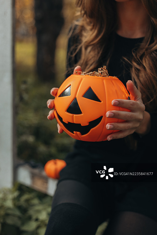 年轻女子坐在栅栏上，手里拿着一个橙色的杰克灯笼水桶，旁边还有一个小南瓜。图片素材