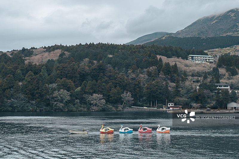日本河口湖上，色彩缤纷的天鹅脚踏船在平静的湖面上悠然滑行，背景是朦胧的森林山峦和湖畔建筑，这是一个阴天秋日的美景。图片素材