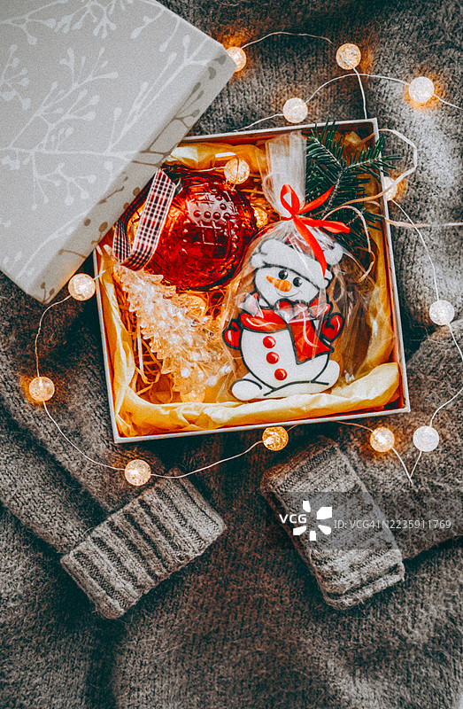 装有红色装饰球的盒子、圣诞树蜡烛、雪人形状的饼干和花环。柔和的灰色毛衣背景增添了温暖感。甜美的节日主题礼品套装。温馨氛围中的圣诞甜点和装饰。图片素材