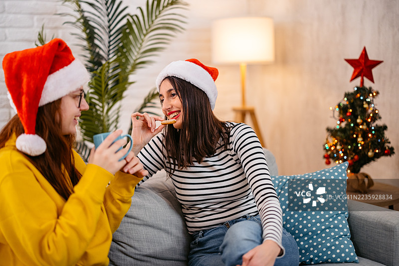 两个女性朋友在圣诞假期期间坐在沙发上喝咖啡、吃饼干并聊天。图片素材