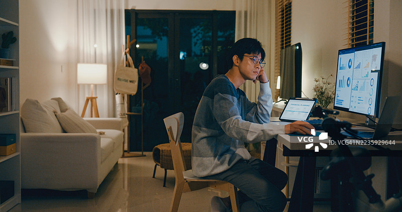 年轻的亚洲男性疲惫的员工在家中办公室加班，因财务规划和数据分析任务感到心理疲惫，面对多台屏幕感到不堪重负。远程工作与财务。图片素材