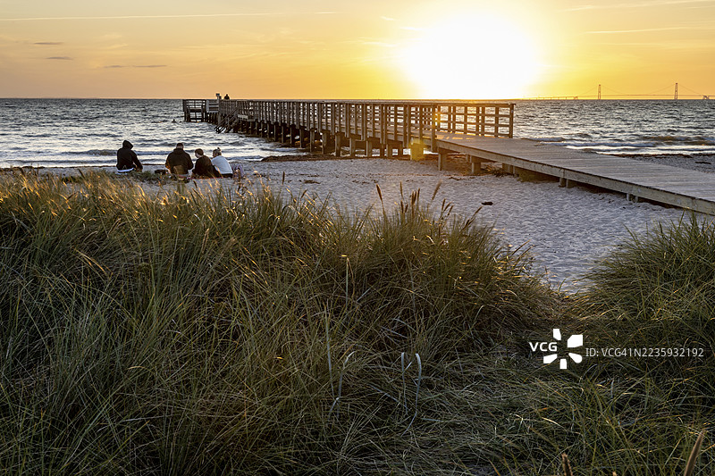 人们坐在海滩上，长长的码头延伸入海，夕阳的光辉映衬着，丹麦西兰岛的科巴克海滩。图片素材