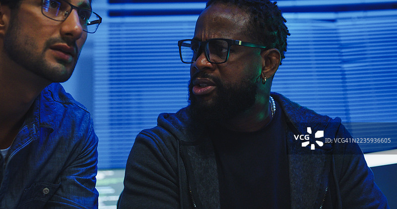 黑人和西班牙裔男性工程师程序员在科技办公室里紧密合作，讨论软件开发策略，直到深夜。软件开发者，人工智能。图片素材