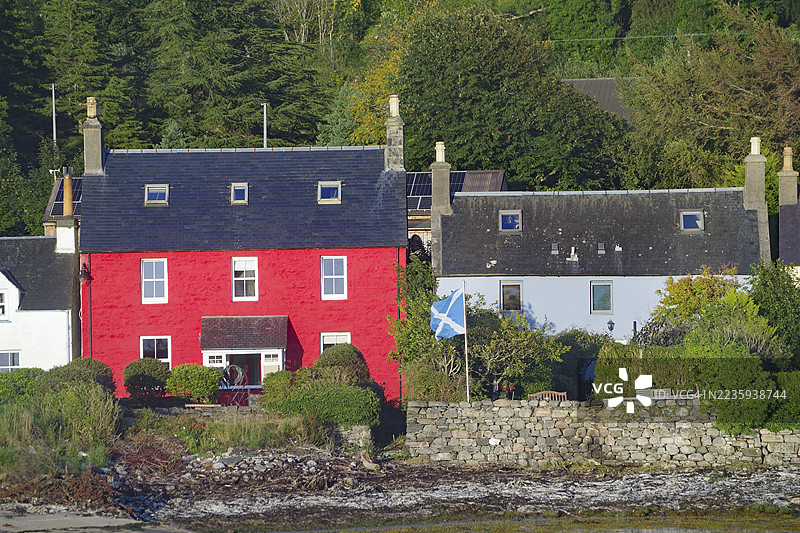 红色房屋上飘扬着苏格兰国旗，四周环绕着树木和石墙，位于海边村庄多尼，苏格兰高地，英国图片素材