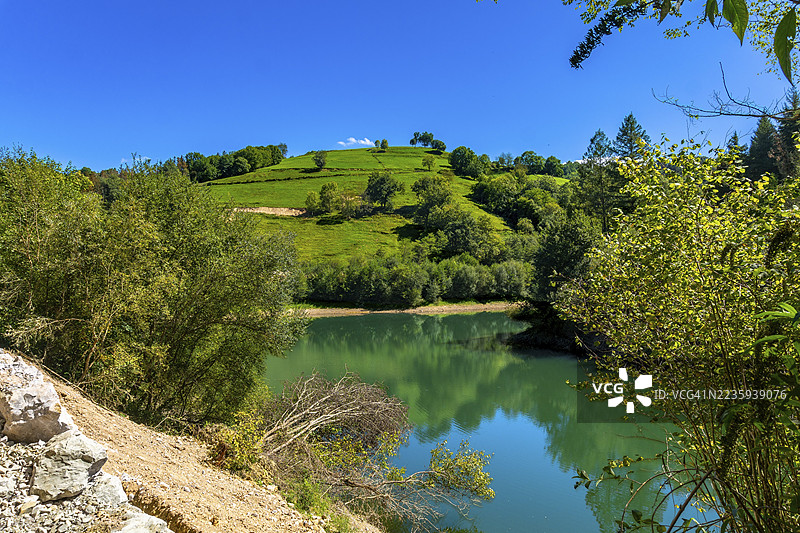 伊比乌尔水库在晴朗的蓝天下映照出周围的绿色山丘和郁郁葱葱的植被，形成了一个宁静而如画的风景，位于吉普斯夸省的托洛萨。图片素材