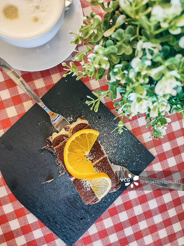 高角度视图的美味巧克力甜点，配有橙子片和一杯咖啡，放在红白格子桌布上。图片素材