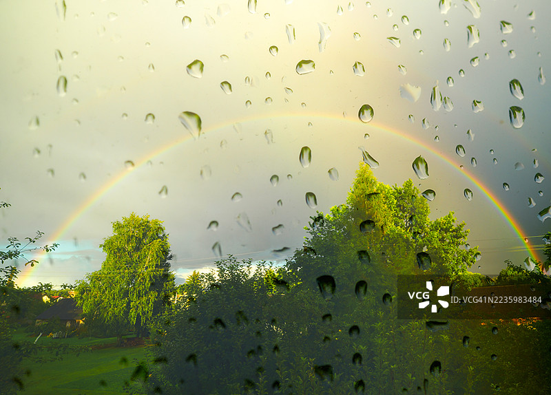 生动的彩虹拱桥横跨郁郁葱葱的绿色景观，前景是沾满雨水的窗户。图片素材