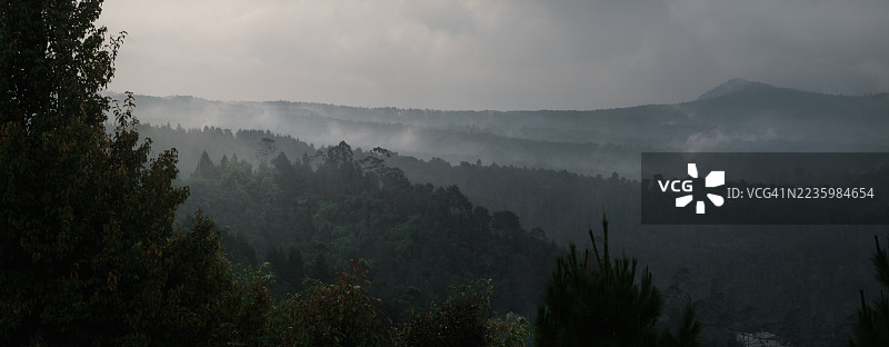 雾气弥漫的山脉和茂密森林在多云的天空下的全景，唤起宁静与神秘的感觉，印度尼西亚图片素材