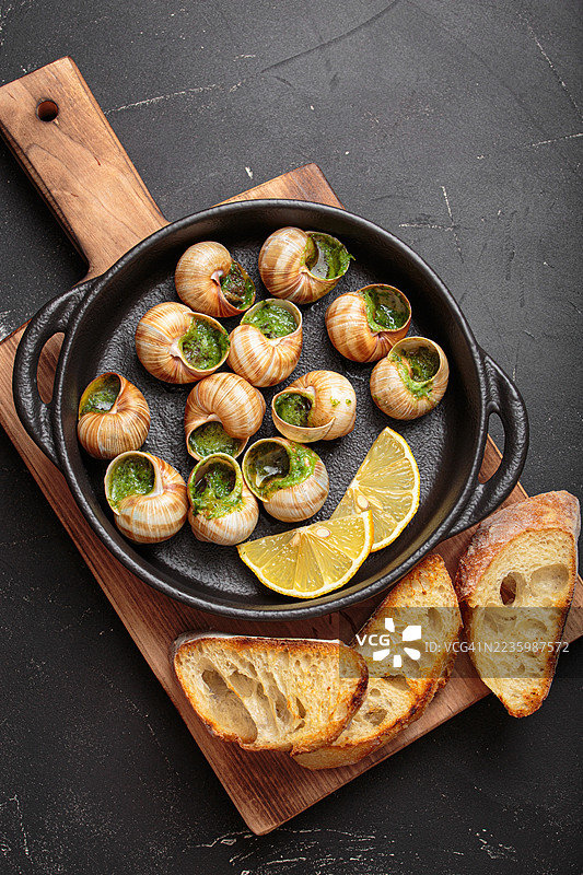 高角度视图的美味法式蜗牛，配有蒜蓉黄油和柠檬，放在铸铁锅中，旁边有烤面包，背景为深色。图片素材