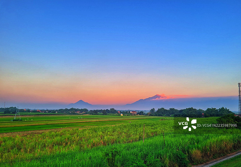 印尼巴东的农业田野上方绚丽的日落，远处有山脉和一座通信塔。图片素材
