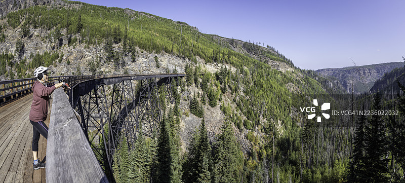 一位年长女性在位于不列颠哥伦比亚省基洛纳的凯特尔谷铁路第6号历史迈拉峡谷高架桥上欣赏秋季郁郁葱葱的森林景色。图片素材