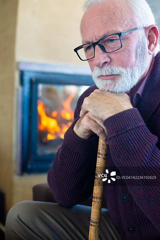 老年男子坐在家中的壁炉前，手持拐杖。图片素材