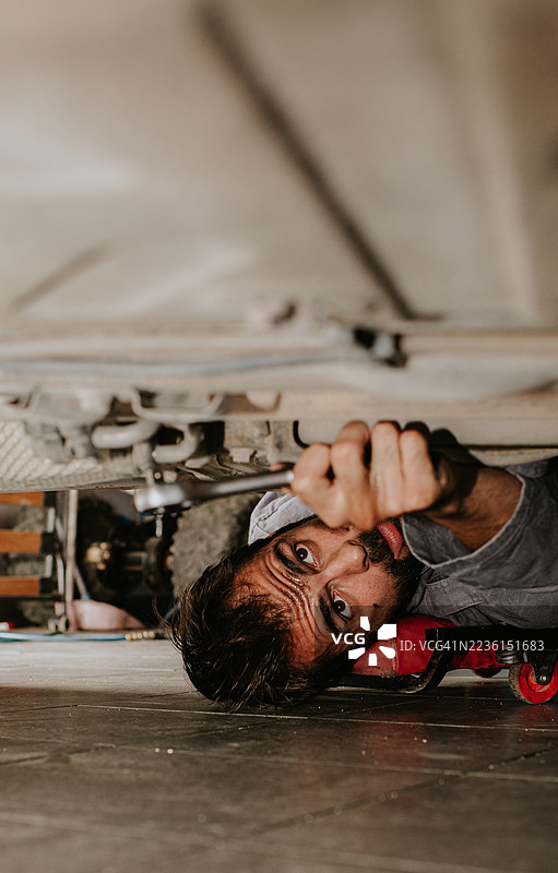 一名机械师在汽车下方滚动，试图检查机械装置。他用扳手伸手到下面，显得很吃力。图片素材