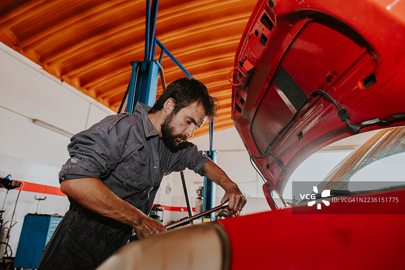 一名穿着工装的机械师正在检查一辆红色汽车的引擎盖。图片素材