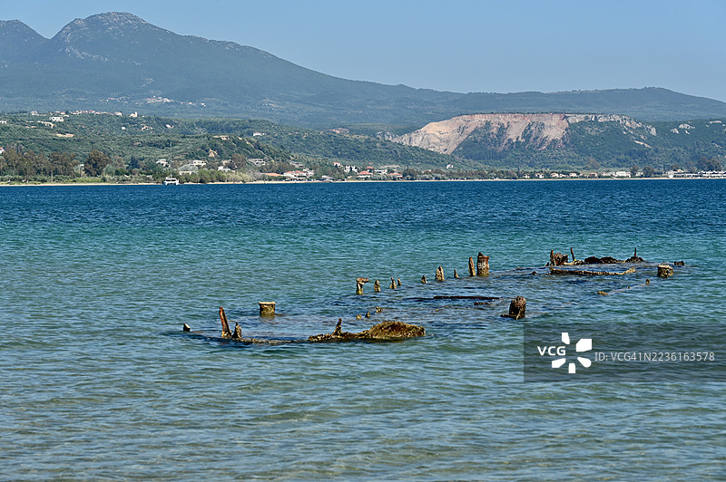 阳光明媚的日子里，希腊清澈蓝水中沉没的船只残骸，背景是山脉海岸线。图片素材
