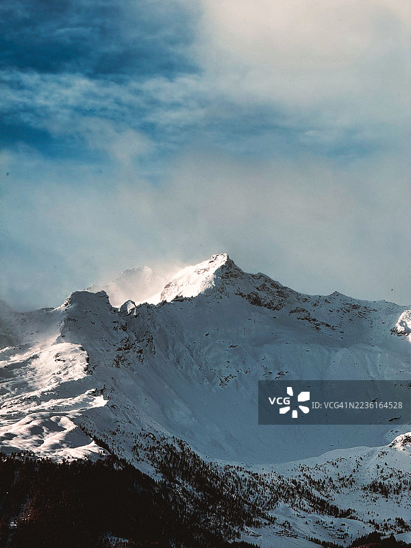 雄伟的雪山峰在戏剧性的天空下被阳光照亮，展现出冬季荒野的崎岖之美，意大利马德西莫图片素材