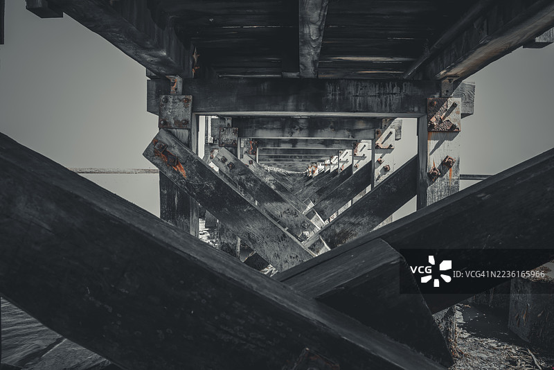 低角度视图的木栈道或桥梁结构，带有风化的横梁和生锈的螺栓，背景是明亮的天空，形成隧道般的透视效果。图片素材