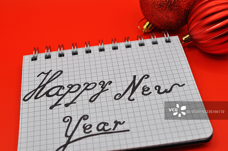 红色背景上，螺旋笔记本上写着“新年快乐”的文字，旁边有红色圣诞装饰品，象征着节日的问候和庆祝。图片素材