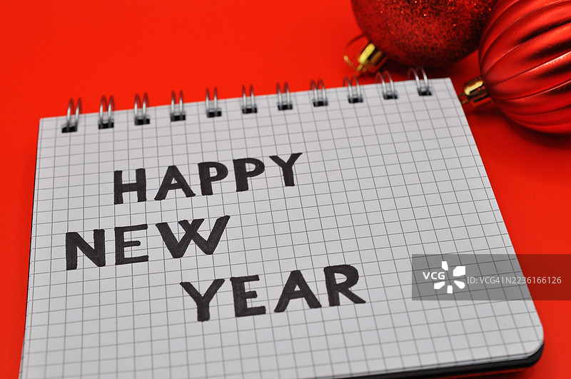 在鲜艳的红色背景上，网格笔记本上写着新年快乐的信息，周围装饰着节日的红色饰品。图片素材