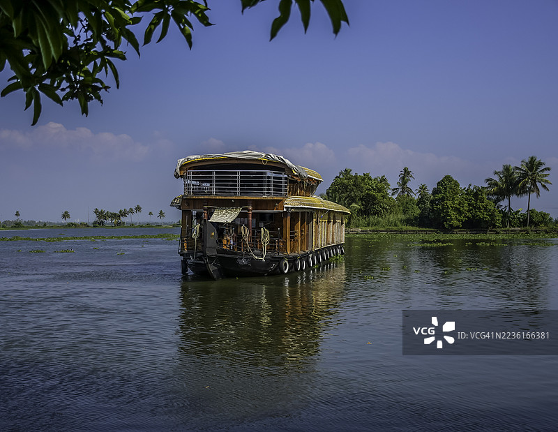 传统木制房船在印度喀拉拉邦宁静的内陆水道上航行，蓝天清澈。图片素材