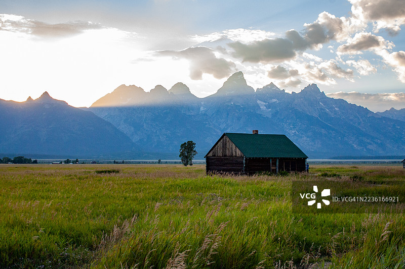 乡村小屋坐落在广阔的草地上，背景是雄伟的山脉和日落时分戏剧性的天空，杰克逊，美国，格兰特顿国家公园图片素材