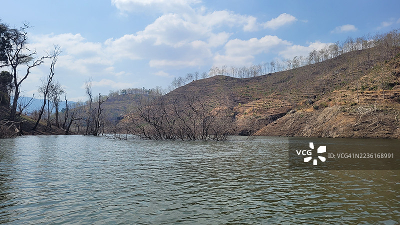 湖泊的美丽景色，光秃的树木和一片光秃的山坡在蓝天和白云下。图片素材