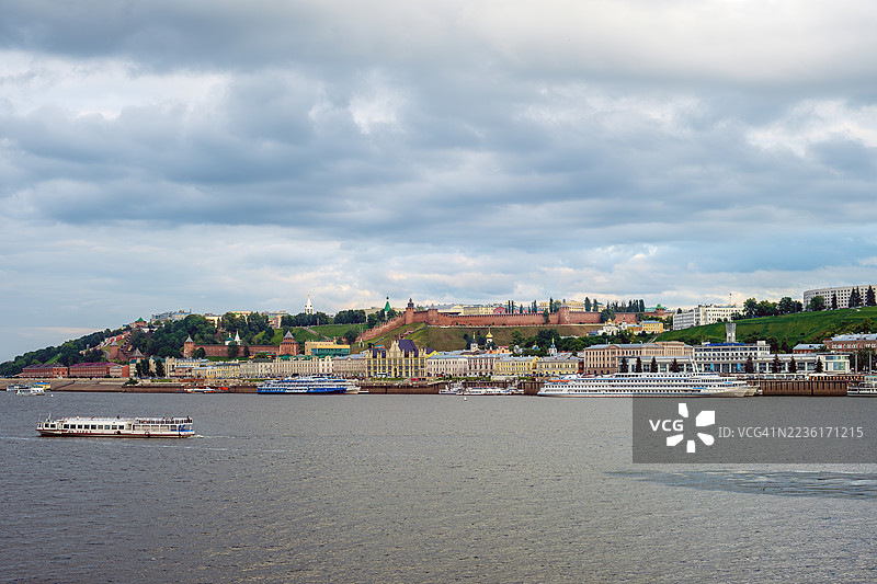 河边山坡上的城市美景，云层密布的天空下，水面上有船只。图片素材