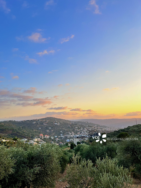 令人惊叹的日落映照在一个坐落于郁郁葱葱的山谷中的风景如画的村庄，前景是橄榄树。图片素材