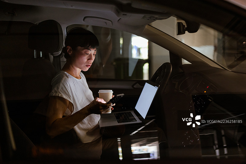 日本女性在车内一边拿着外卖咖啡，一边用手机滑动，旁边有笔记本电脑。图片素材