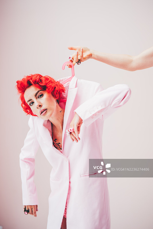 一个留着醒目红发的模特，身穿oversize粉色西装外套，手持衣架，被挂在粉色衣架上。图片素材