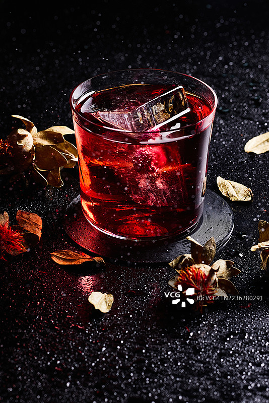 深色背景上，一杯加了冰块的内格罗尼鸡尾酒，周围散落着干枯的树叶。图片素材