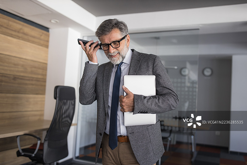 一位年长的商务人士在办公室里一边接电话一边拿着笔记本电脑图片素材