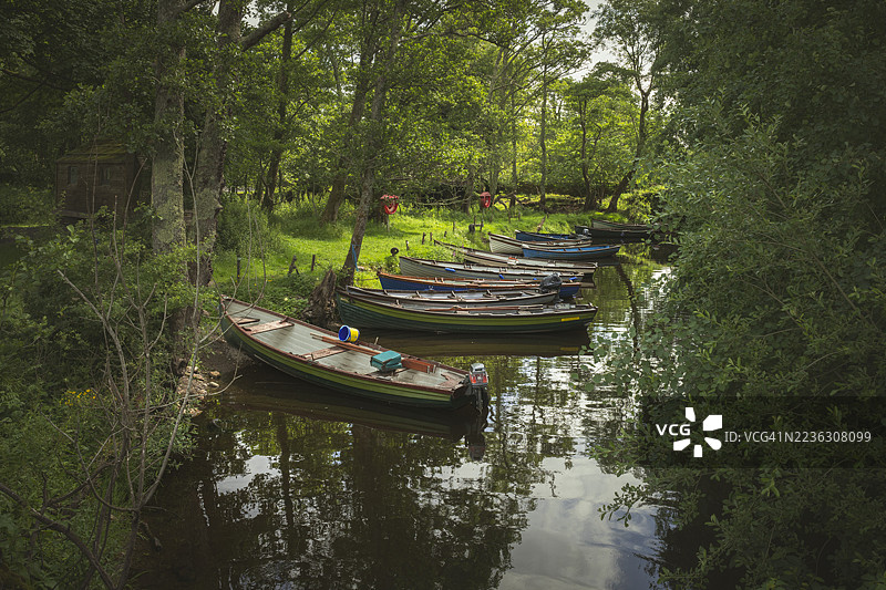 爱尔兰基拉尼国家公园，色彩斑斓的船只沿岸停泊在郁郁葱葱的绿色河岸边，倒映在平静的水面中，俯瞰视角。图片素材