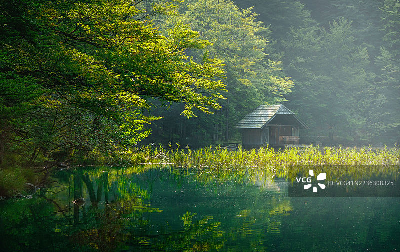 宁静的木屋坐落于森林湖畔，伴随着日出，郁郁葱葱的绿色植被与水面倒影交相辉映，奥地利，朗巴特湖图片素材
