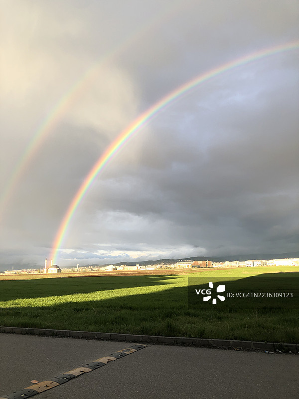 双彩虹拱形横跨绿色田野和远处城市，背景是戏剧性的天空图片素材