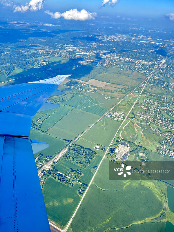 蓝天白云下，前景是飞机机翼，俯瞰绿色农业田地和郊区景观。图片素材