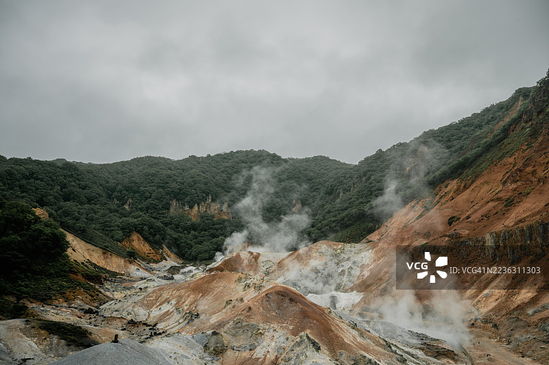 日本地狱谷展望台，多云天空下，蒸汽从地热喷口升起，周围是郁郁葱葱的绿色山脉，构成戏剧性的火山地貌图片素材