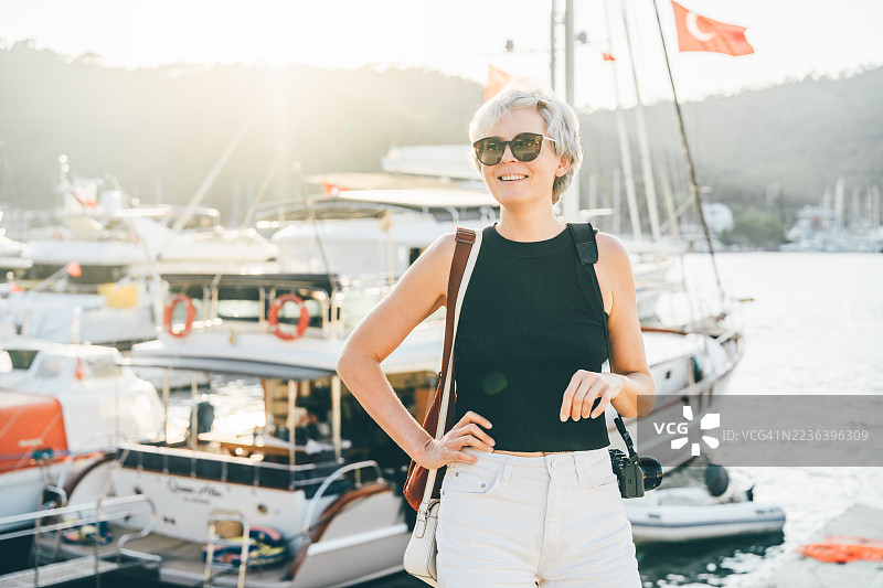 一位快乐的成熟的女性，短短的灰色头发和太阳镜，微笑着在码头上摆姿势，背景是游艇。图片素材